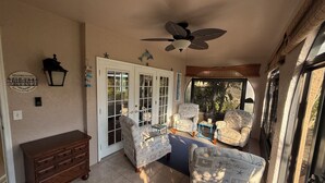 A sunlit room with a ceiling fan, two patterned armchairs, a wooden dresser, and a sliding glass door leading outside.
