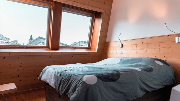 A bedroom with a wooden sloped ceiling, a bed with a patterned bedspread, a bedside table, and a window with a view of buildings and trees. - Immodreams - La Patinoire - Avoriaz (Avoriaz)