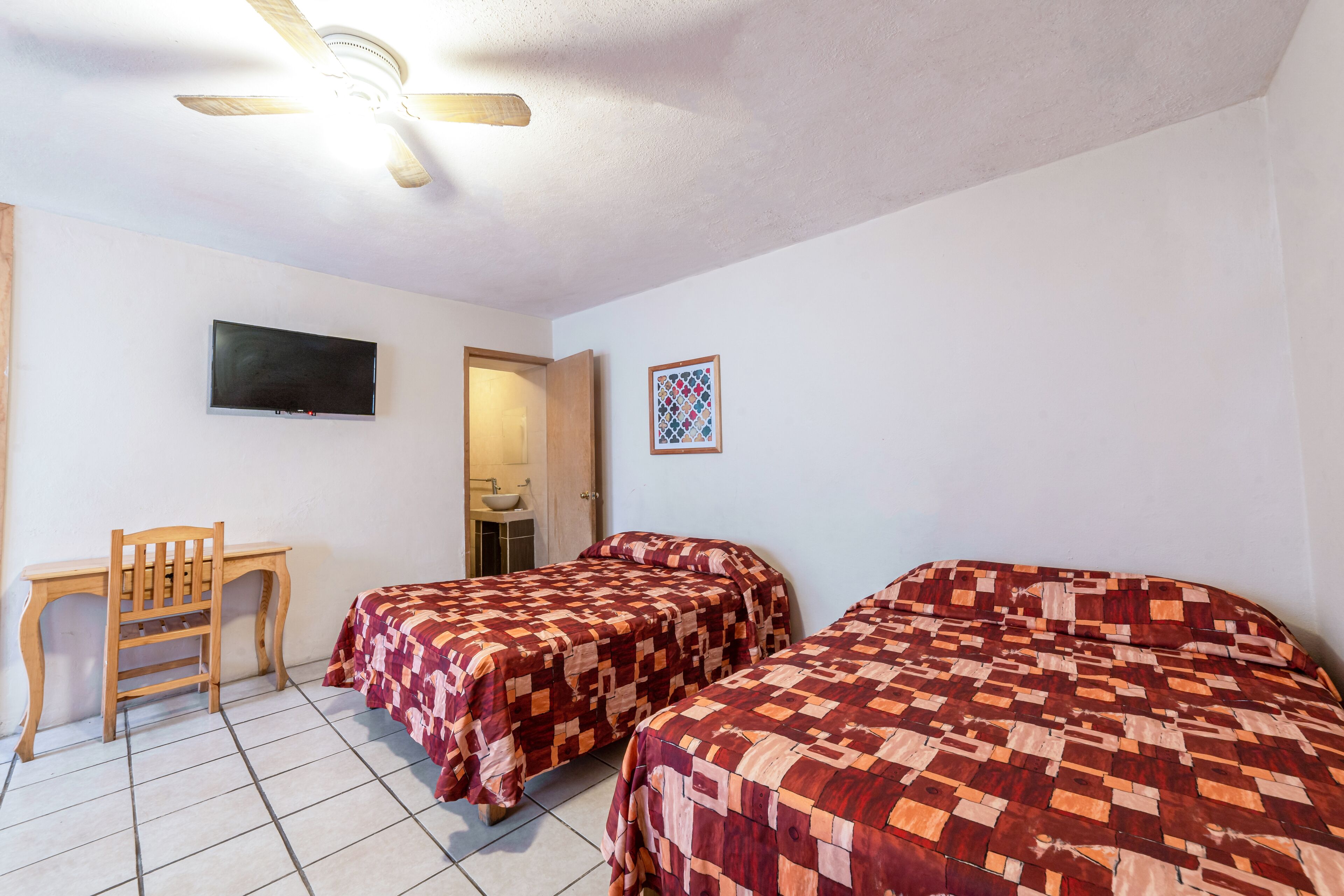 Una habitación con dos camas, un escritorio, una silla, un ventilador de techo y un televisor en la pared.