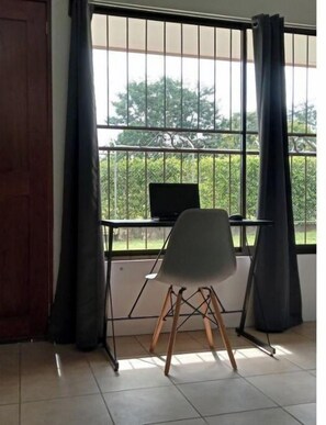 Habitación con escritorio y silla cerca de una ventana con barrotes, cortinas y vistas al exterior con vegetación.