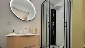 A modern bathroom with a glass-enclosed shower, a round mirror, and a vanity with toiletries. - Chambre LES Pins (Mimizan)