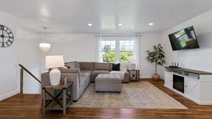 Un salón amplio con un sofá modular, una mesita con una lámpara, un televisor montado en la pared y una escalera que sube a otro nivel.