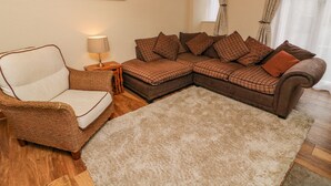 A living room with a brown sectional sofa, a wicker armchair, a small side table with a lamp, and a large beige rug. - No. 2 The Mews (Carnforth)