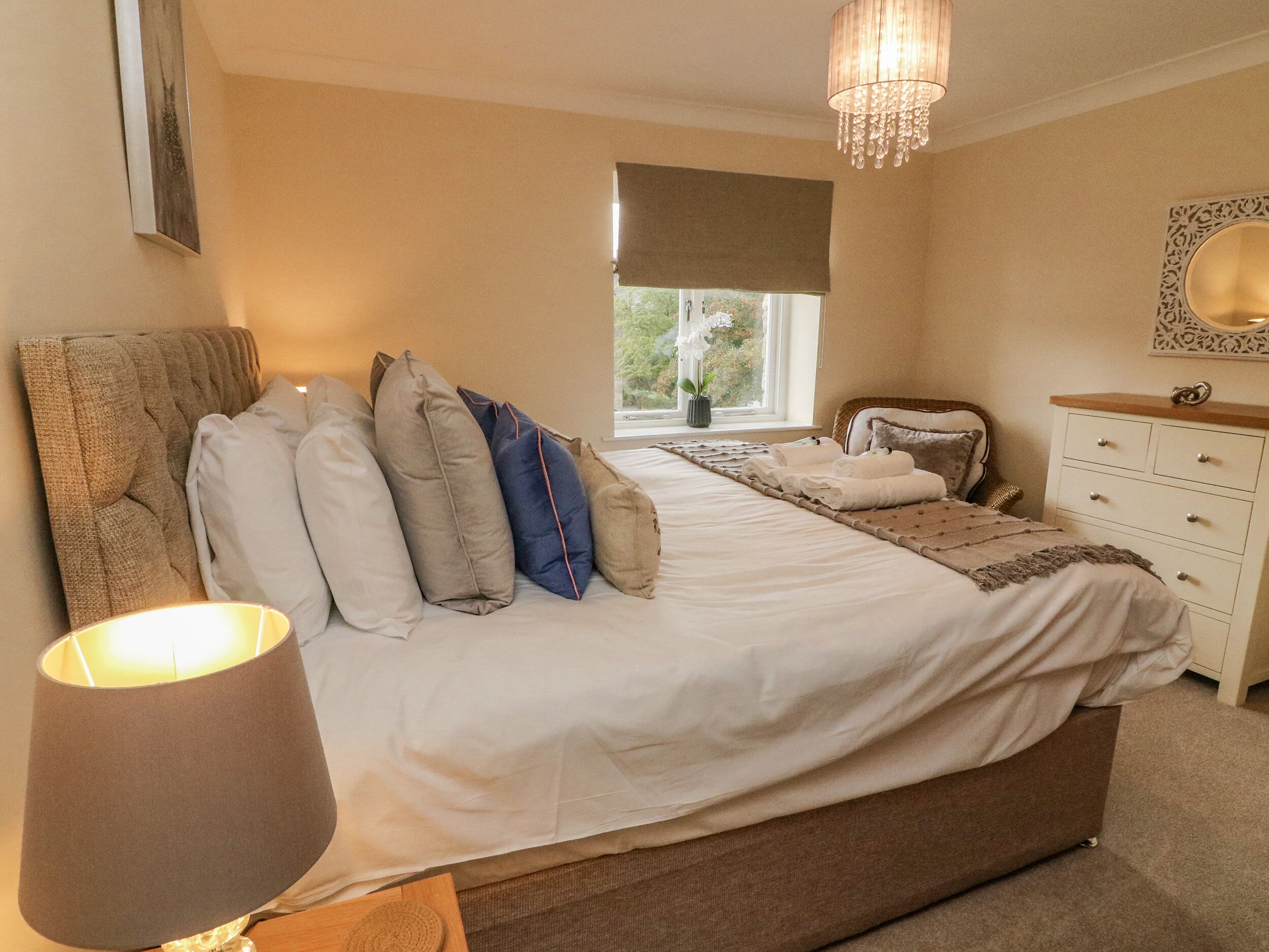 A bedroom with a large bed, a bedside table, a dresser, a window with a view, and a chandelier.