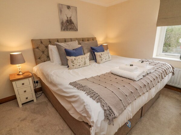 A neatly made bed with a beige headboard, white bedding, and a decorative throw blanket. There is a bedside table with a lamp and a small drawer. A framed artwork hangs above the bed.