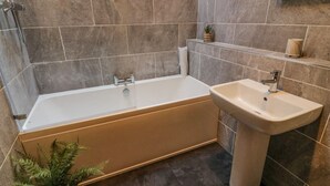 A modern bathroom with a bathtub and a pedestal sink, tiled walls, and a small shelf with toiletries. - No. 2 The Mews (Carnforth)