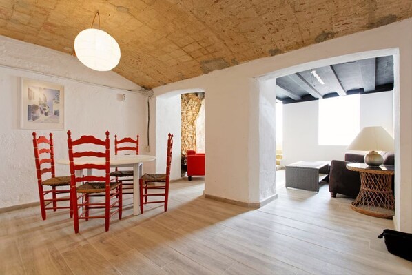 A spacious room with a wooden floor, a dining area with red chairs, a white table, and a large window.