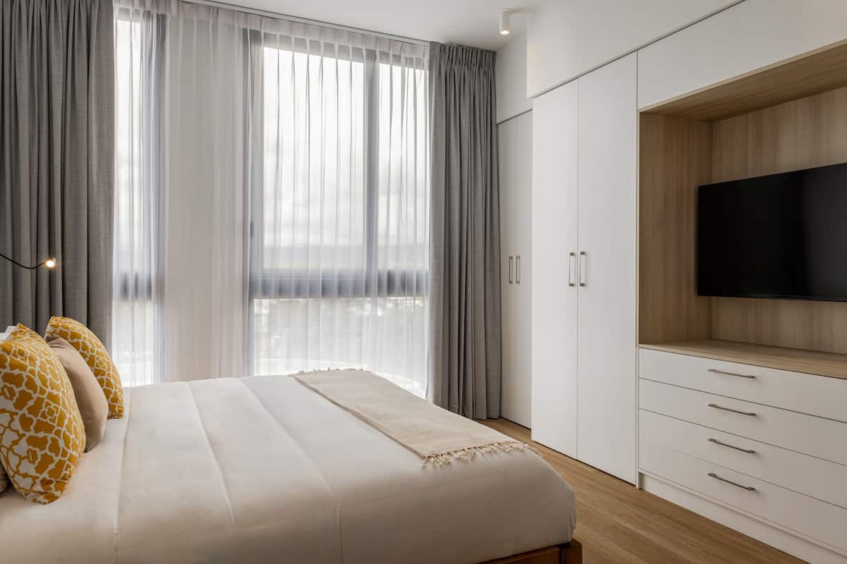 A bedroom with a large bed, a TV mounted on a wooden cabinet, and a view of the city through sheer curtains.