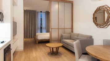A modern hotel room with a wooden floor, a sofa, a round wooden table, and a bed with curtains.