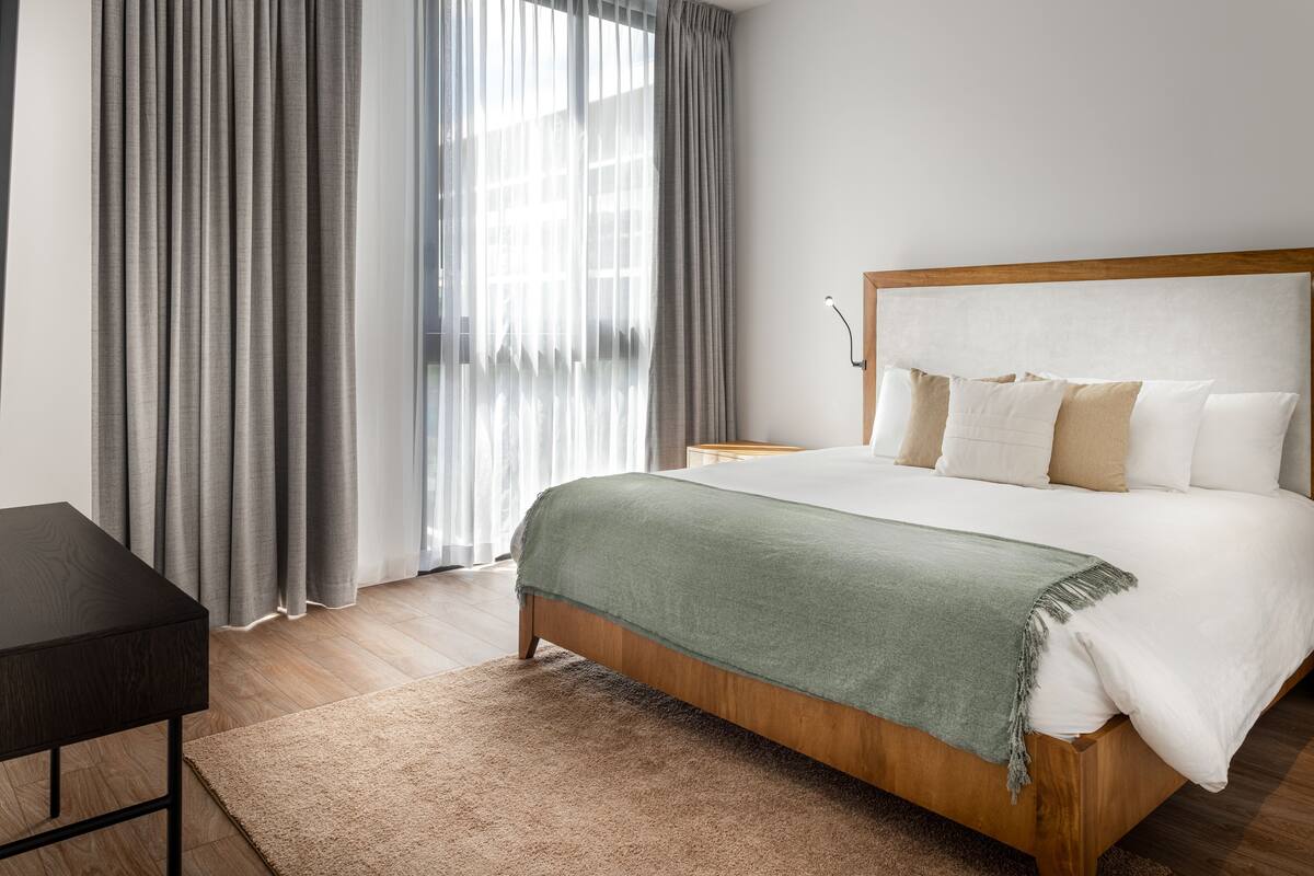 A bedroom with a wooden bed, a bedside table, a lamp, and a large window with curtains.