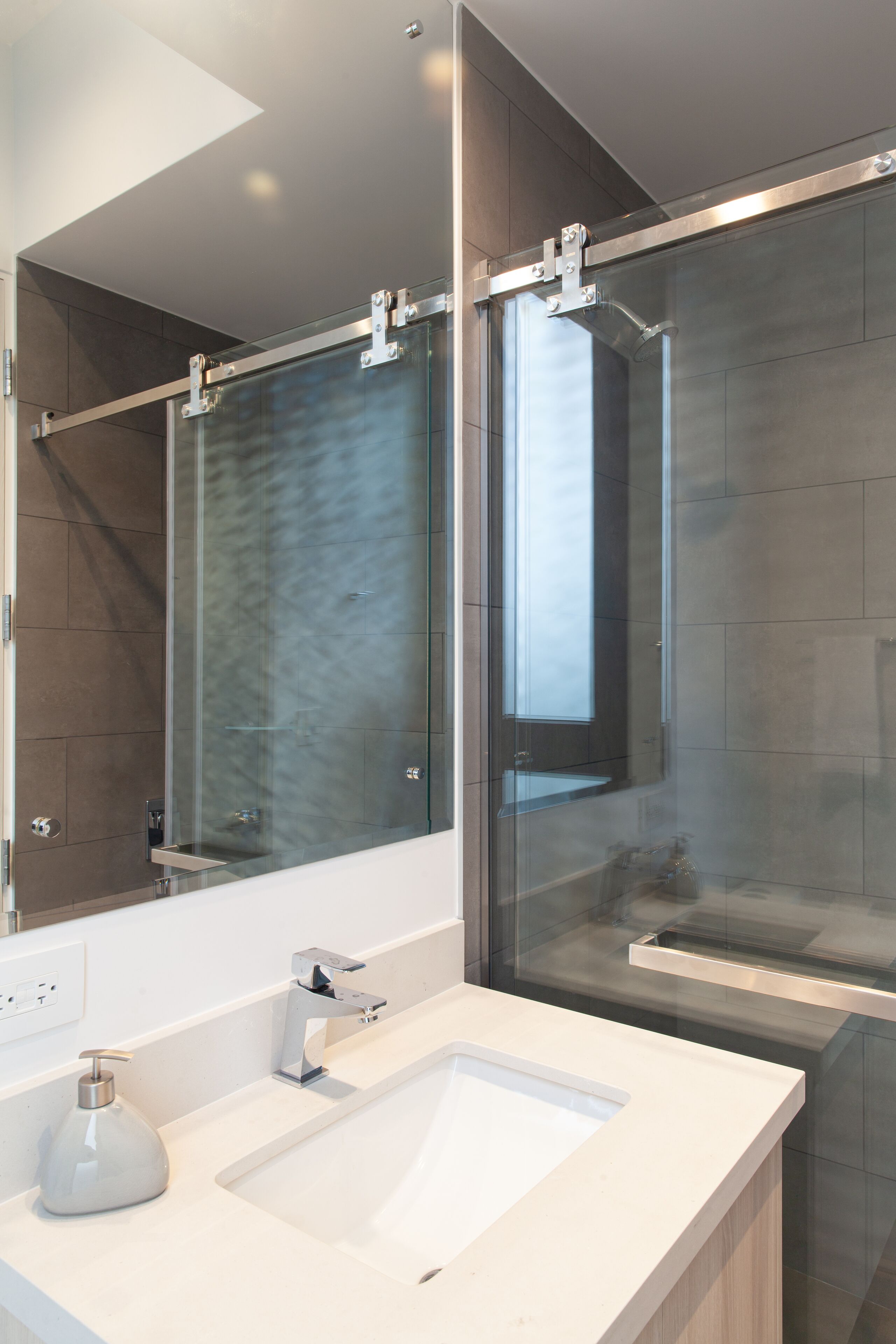 Une salle de bain moderne avec une douche à parois de verre, un grand miroir et un lavabo blanc doté d’un distributeur de savon.