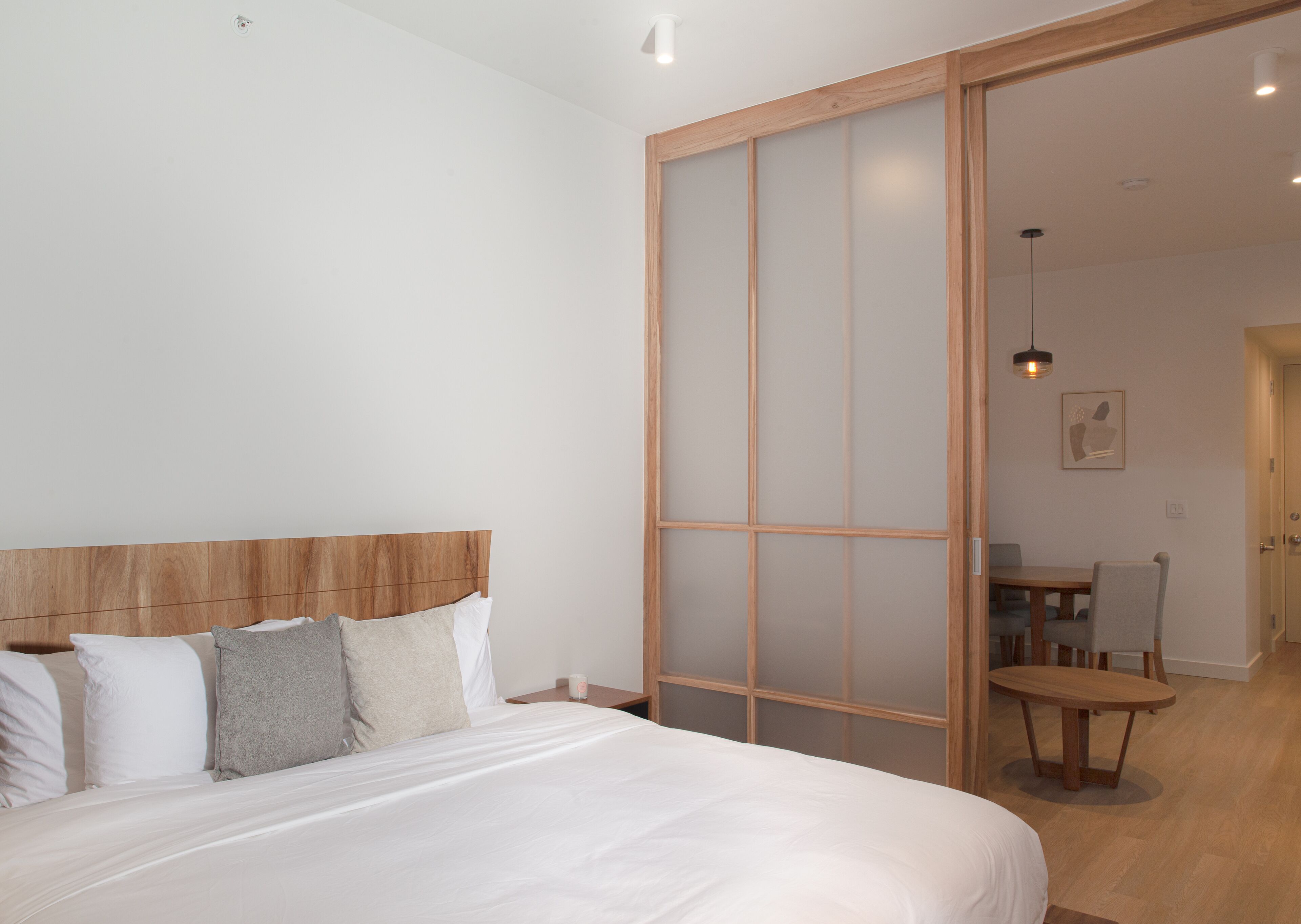 Quarto com cabeceira de madeira, cama com roupa de cama branca e porta de vidro deslizante para uma área de jantar com mesa e cadeiras.