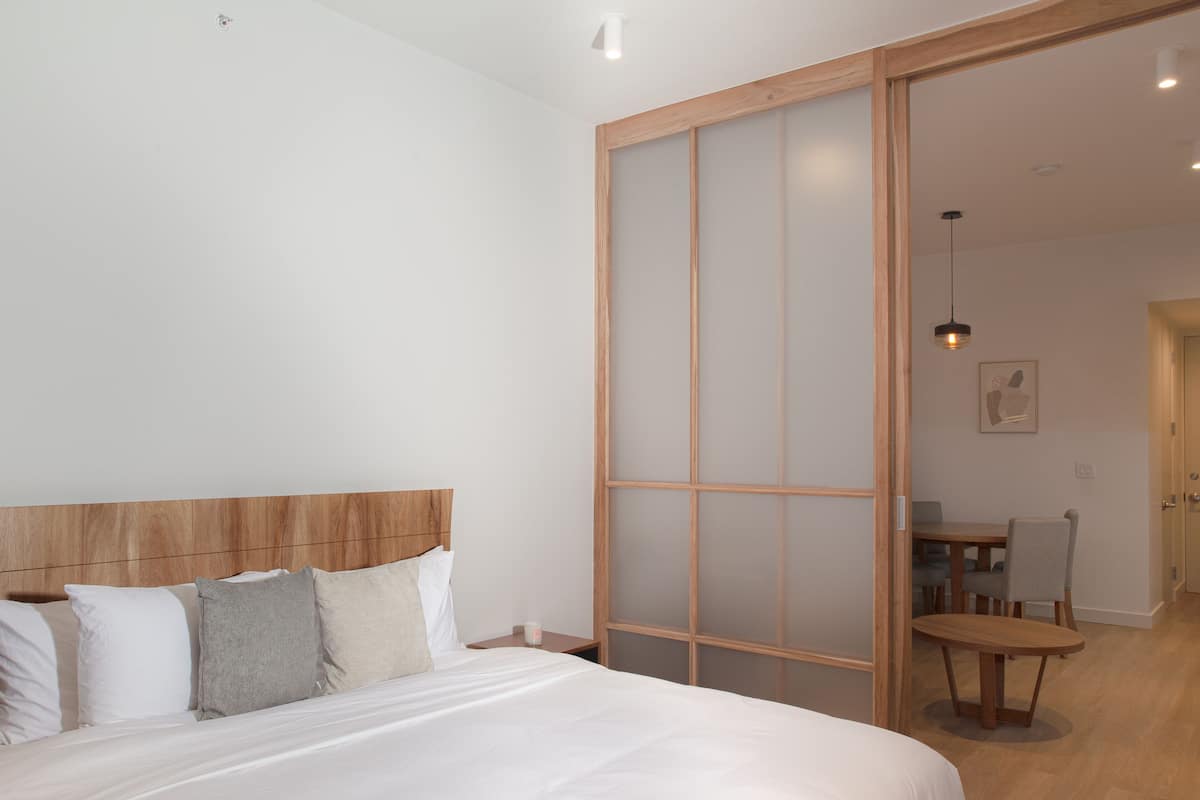 A bedroom with a wooden headboard, a bed with white bedding, a sliding glass door leading to a dining area with a table and chairs.