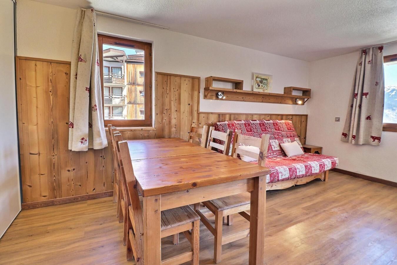 A wooden dining table with chairs, a wooden sofa with patterned cushions, and a window with curtains.