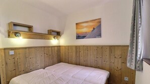 Una habitación con cabecera tapizada de madera, una cama con edredón blanco, un cuadro enmarcado de un paisaje de montaña y una pequeña estantería con una lámpara.