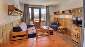 Una habitación acogedora con un sofá de madera, una mesita, un televisor y vistas a montañas a través de amplios ventanales.