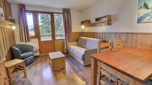 A cozy room with a wooden floor, a sofa, a dining table, and a chair. There are curtains on the windows and a painting on the wall.