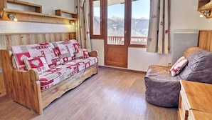 Une pièce chaleureuse avec un canapé en bois et un banc rembourré, une étagère en bois avec une lampe, et une fenêtre donnant sur les montagnes.