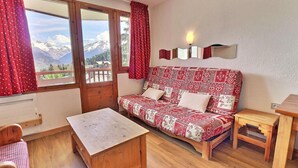 A cozy room with a wooden sofa, a small wooden table, and a view of mountains through large windows. - 10m from ski slopes, 7th floor, balcony, ski locker, 25m², La Tania (Courchevel)