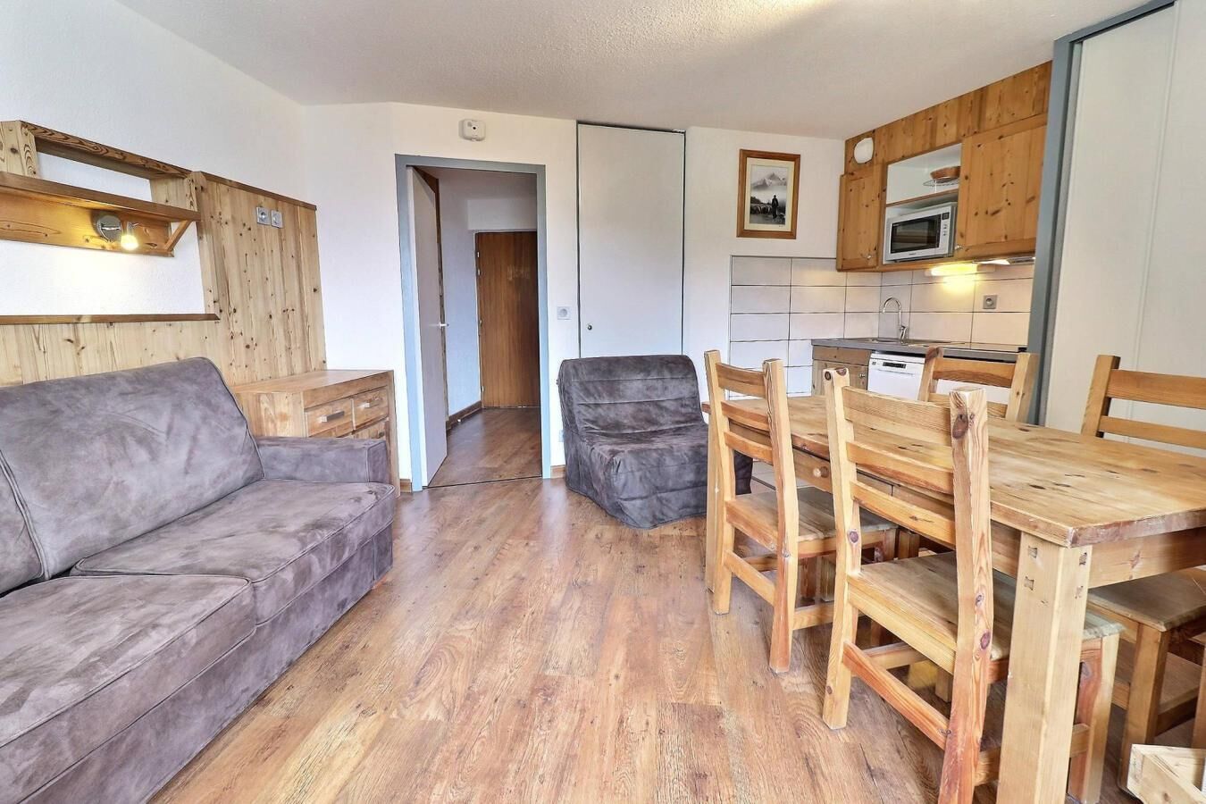 Een gezellige woonkamer met een grijze bank, een houten eettafel met stoelen, en een keuken met houten kastjes en een magnetron.