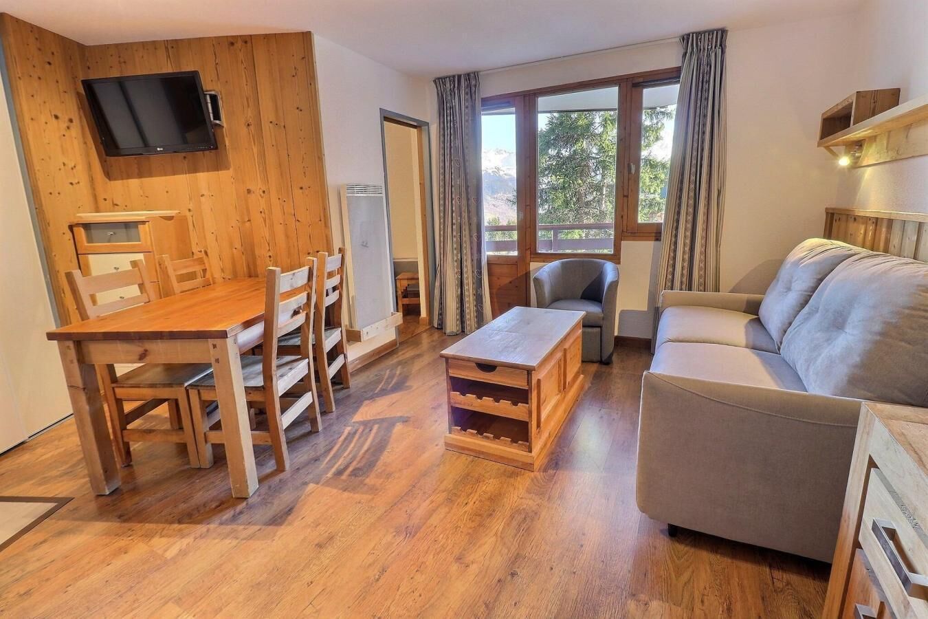 Ein Wohnzimmer mit Holzboden, einem Esstisch aus Holz, einer Couch und einem an der Wand befestigten Fernseher.