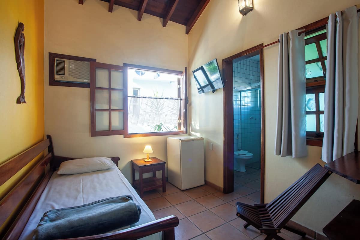 Una habitación compacta con cama, una mesita de noche con lámpara, un refrigerador y un banco.