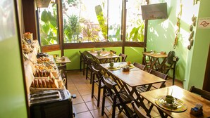 Un restaurante con varias mesas y sillas de madera, una zona de bufé con diferentes platillos, y ventanas amplias que ofrecen vistas a la vegetación exterior.