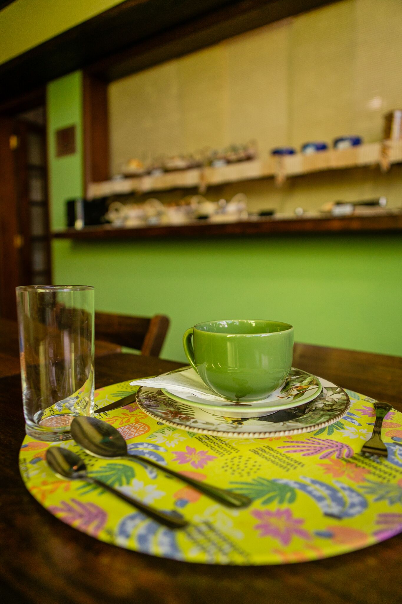 A dining table with a green cup and saucer, a glass, and cutlery on a colorful placemat.