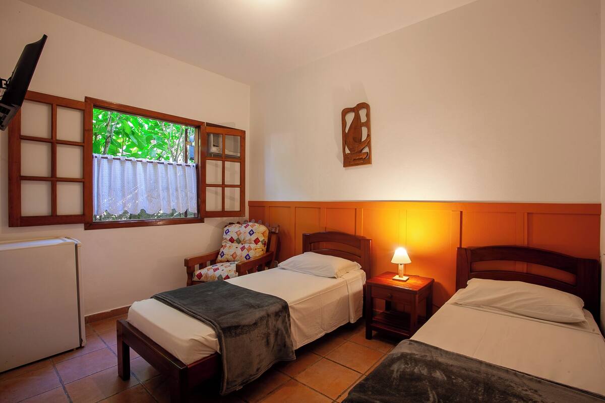 Una habitación con dos camas individuales, una mesita de noche con lámpara, una silla, una ventana con vistas a zonas verdes y un radiador.