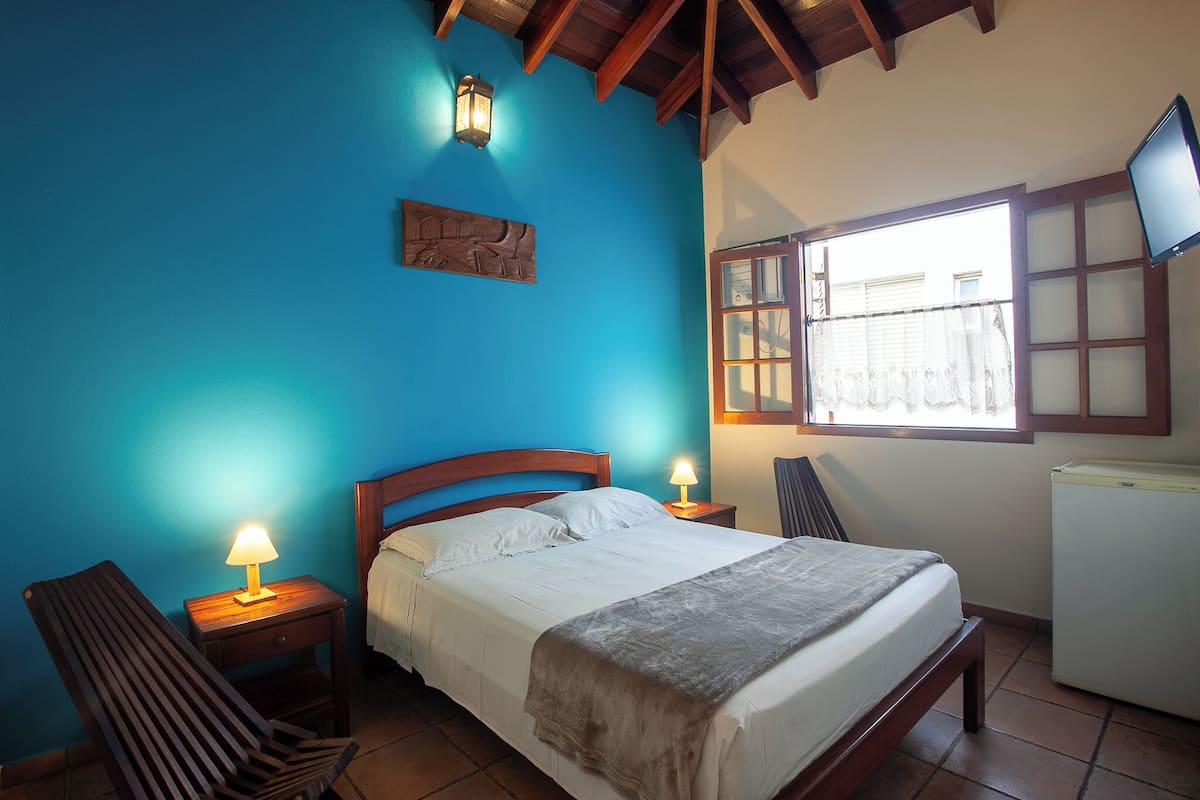 Un dormitorio con cama, dos mesitas de noche con lámparas, una silla de madera, una ventana con cortinas y un televisor montado en la pared.