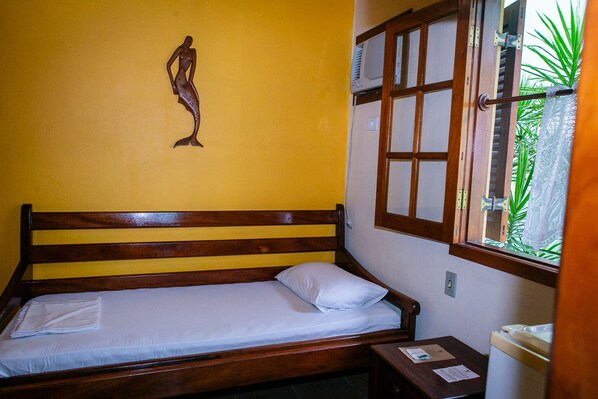Una cama individual con cabecera de madera y colchón blanco, una mesita de noche de madera con lámpara, y una ventana con vista a la vegetación.
