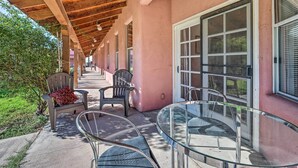 A covered outdoor seating area with chairs and a glass table, adjacent to a building with large windows and a pink exterior.