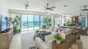 A spacious living area with a large sofa, a wooden coffee table, and a TV on a cabinet. There are ceiling fans, large windows with a view of the ocean, and a kitchen area visible in the background. - Bakers Bay: Key Largo Sunset Haven (Key Largo)