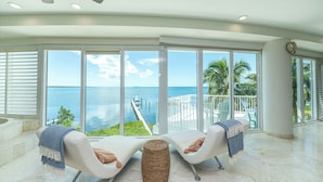 A spacious room with two white lounge chairs, a woven stool, and large windows offering a view of the sea and a dock. - Bakers Bay: Key Largo Sunset Haven (Key Largo)