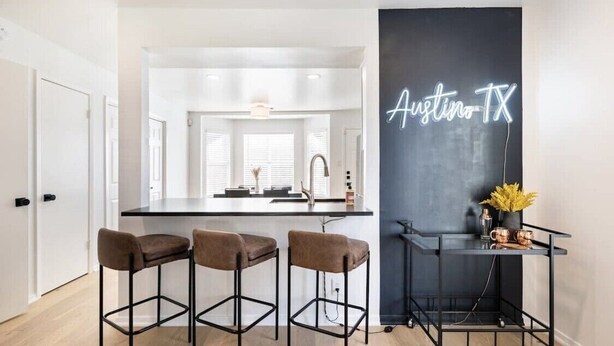 A modern kitchen with a bar area, a neon sign reading "Austin TX", and a cart with mugs. - Spacious 2 Bedroom East Riverside Home with Garage and Yard (Austin)