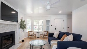 A modern living room with a fireplace, a large flat-screen TV, a dark blue sofa, two wicker chairs, a glass coffee table, and a ceiling fan. - Spacious 2 Bedroom East Riverside Home with Garage and Yard (Austin)