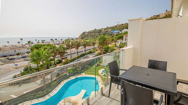 A balcony with a glass railing overlooking a swimming pool, a beach, and a road with palm trees. - Paloma Beach apartment 3.2a (Manilva)