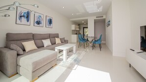 A modern living room with a grey sofa, a white coffee table, and blue chairs. There are abstract paintings on the wall and a TV on the right. - Paloma Beach apartment 3.2a (Manilva)