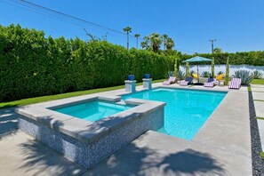 Una piscina rectangular con borde de azulejos, rodeada por un seto, y un pabellón con sillas al fondo.