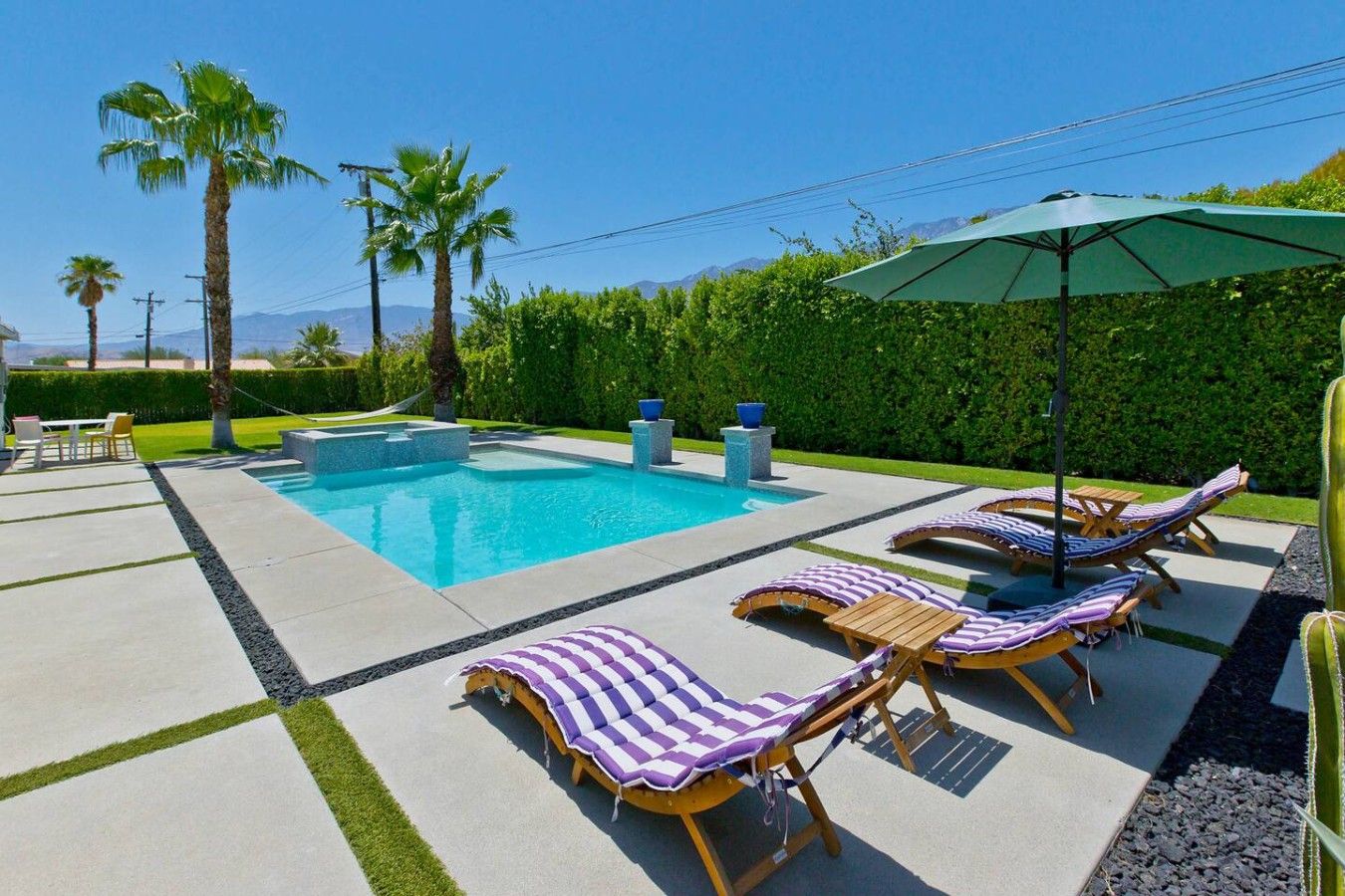 A swimming pool with lounge chairs and an umbrella, surrounded by palm trees and a hedge.