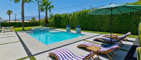 A swimming pool with lounge chairs and an umbrella, surrounded by palm trees and a hedge.