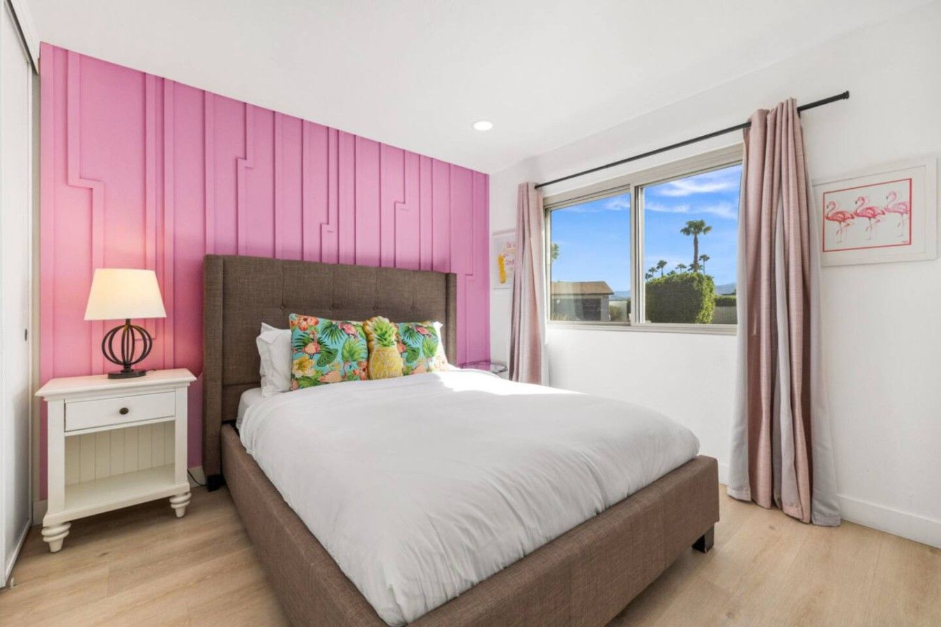 Une chambre à coucher comprenant un lit, une table de chevet, une lampe, une fenêtre avec des rideaux et un mur orné d’un panneau décoratif rose.
