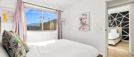 A bedroom with a large bed, a window with curtains, a framed picture, and a view of the outdoors.