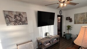 Een woonkamer met een houten vloer, een plafondventilator, een flatscreen televisie, een bijzettafeltje met boeken, een lamp en een schilderij aan de muur.