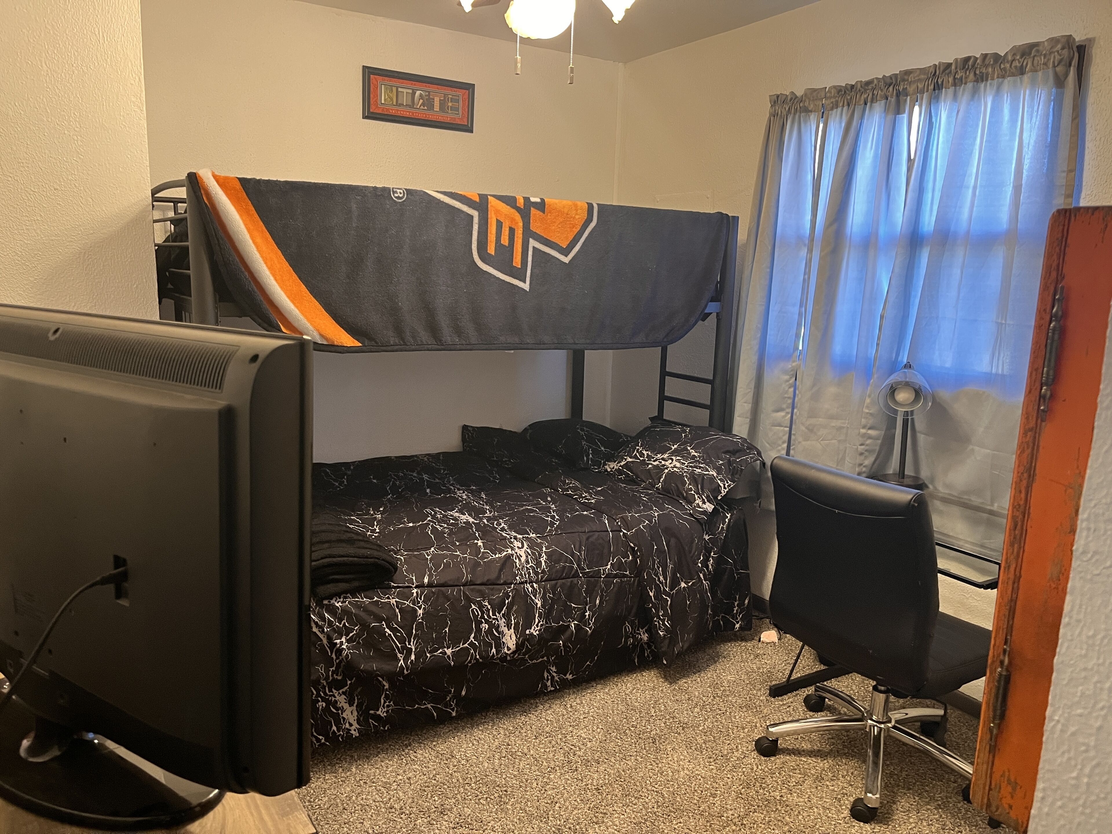 Una cama individual en literas con el logotipo de un equipo deportivo, un escritorio con una computadora, una silla y una ventana con cortinas.
