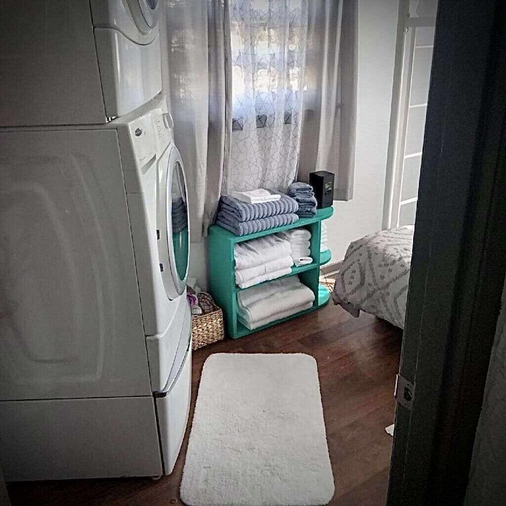 A laundry area with a washing machine, a shelf with folded towels, and a bed with a patterned bedspread.