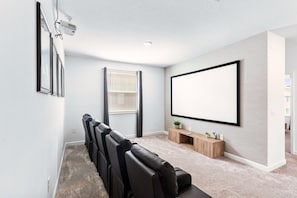 A home theater room with a large flat-screen TV, a projector, and a row of black leather chairs. - Vacation 6BR Home w Pool Spa & Patio (Kissimmee)