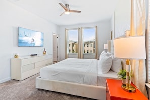 Une chambre à coucher comprenant un lit, une table de chevet avec une lampe, une commode, un ventilateur de plafond et une fenêtre avec des stores.