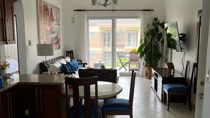 A spacious living area with a dining table, chairs, and a sofa. There is a kitchen area with cabinets and a water dispenser. A large window provides a view of the outside.