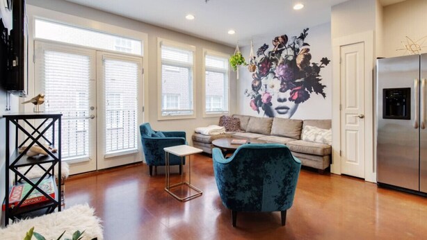 A modern living room with a sofa, two armchairs, a coffee table, and a TV. There are large windows with blinds, a refrigerator, and a decorative wall piece.